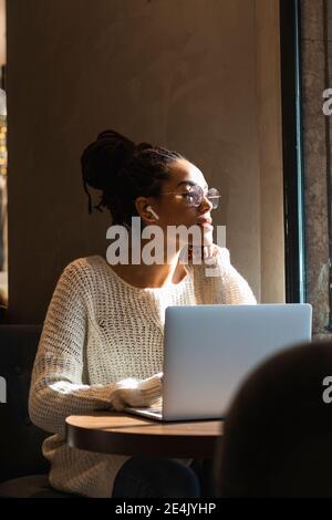 Giovane donna pensierosa che guarda via mentre si siede con il computer portatile a. caffè Foto Stock