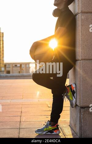 Sole che splende attraverso il giovane uomo che tiene il basket mentre si sta in piedi muro al tramonto Foto Stock