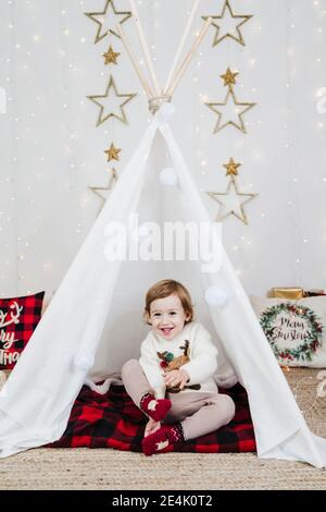 Allegra ragazza cute del bambino seduta in tenda contro le decorazioni di Natale a casa