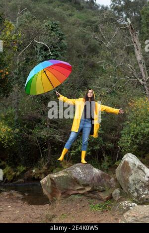 Giovane donna spensierata che indossa un impermeabile giallo che tiene l'ombrello mentre si è in piedi su roccia nella foresta Foto Stock