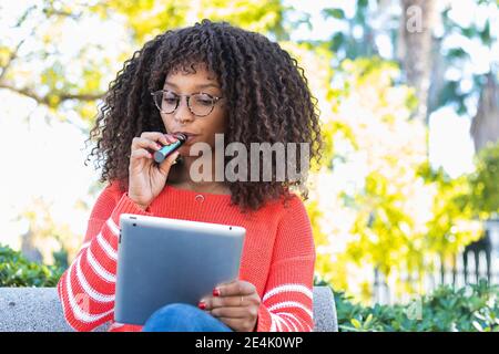 Donna afro fumando sigaretta elettronica mentre si usa tablet digitale in parco pubblico Foto Stock