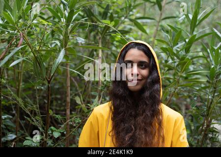 Giovane donna che indossa impermeabile giallo contemplando mentre si sta in piedi contro le piante in foresta Foto Stock