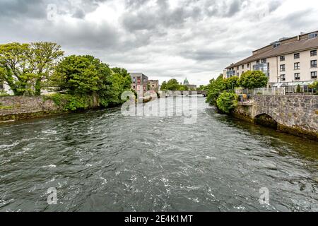 Fiume Corrib tra pareti in pietra e vegetazione verde con il ponte William o'Brien e la cupola verde della Cattedrale sullo sfondo, nuvoloso da Foto Stock