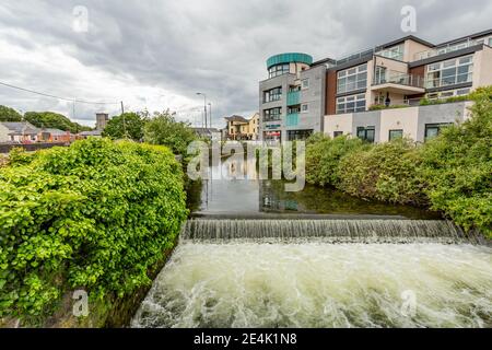 Galway, provincia di Connacht, Irlanda. 11 giugno 2019. Fiume medio che corre lungo il fiume Corrib, piccola diga che forma una cascata, edifici nel backgr Foto Stock