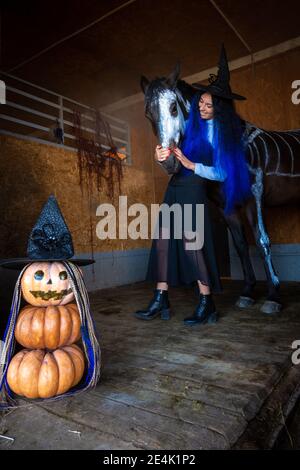 Una ragazza in costume di strega abbraccia un cavallo in un Corral, in primo piano una figura malvagia di zucche Foto Stock