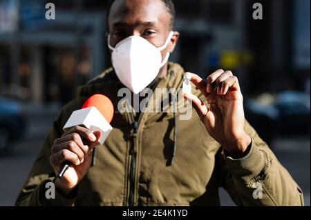 Primo piano di reporter maschile indossando maschera facciale che mostra il vaccino mentre parlare con il microfono Foto Stock