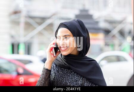 Ritratto di giovane bella donna con hijab nero che parla con smartphone in mezzo alla strada Foto Stock