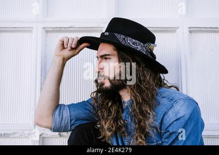 Elegante giovane uomo con un look cool che tiene cappello contro il bianco parete Foto Stock