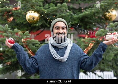 Giovane sorridente che tiene dono e ornamento contro l'albero di Natale Foto Stock