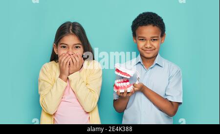 Bambini multietnici spaventati che mostrano mal di denti su sfondo turchese. Ragazza che nasconde la bocca dietro le mani e ragazzo che tiene il modello mascellare nelle mani Foto Stock