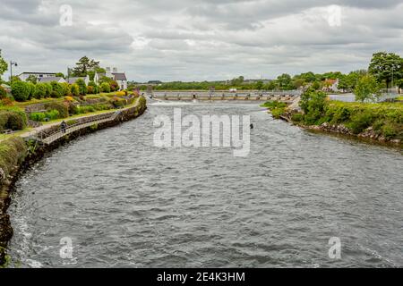 Galway, provincia di Connacht, Irlanda. 11 giugno 2019. Acqua che scorre sul fiume Corrib verso Salmon Weir tra la vegetazione verde, con due pescatori, Foto Stock