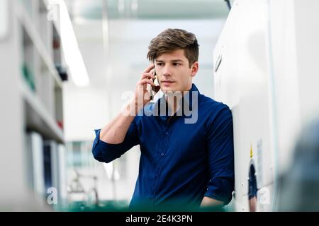 Ingegnere maschile che parla di chiamata mentre si appoggia al macchinario attrezzatura in fabbrica Foto Stock