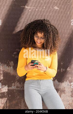 Giovane donna alla moda che usa il telefono cellulare mentre si appoggia alla parete Foto Stock