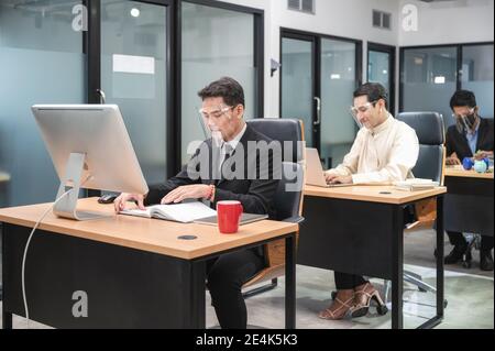 Dipendente asiatico che indossa la visiera che lavora su una scrivania in legno con desktop in un nuovo ufficio normale in uno spazio di lavoro Foto Stock