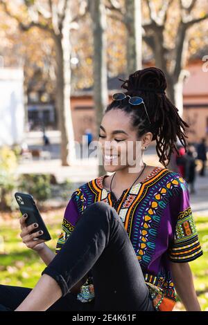 Sorridente giovane donna alla moda che usa lo smartphone mentre si è seduti a. parcheggio Foto Stock