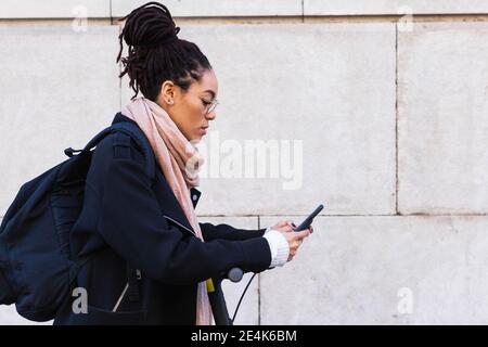 Donna giamaicana con Dreadlock utilizzando smartphone mentre si appoggia spingere lo scooter a parete Foto Stock