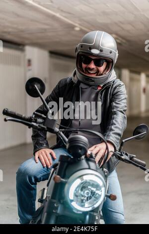 Uomo allegro che indossa casco e occhiali da sole mentre si siede in moto nel parcheggio Foto Stock