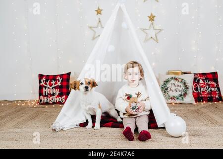 Carina bambina con cucciolo seduto in tenda contro Natale decorazioni a casa