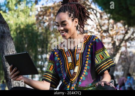Sorridente bella donna giamaicana con Dreadlock utilizzando tablet digitale AT parcheggia il giorno di sole Foto Stock