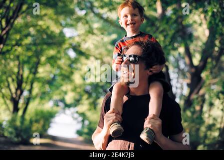 Il giovane padre felice tiene il suo figlio sulle spalle e passeggiate in estate nel parco cittadino Foto Stock