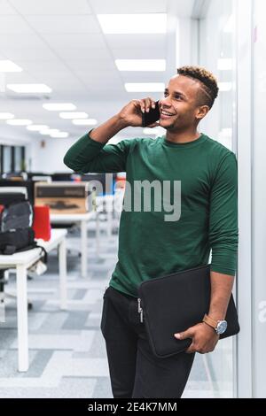 Uomo d'affari sorridente che tiene il documento di file mentre parla durante la chiamata telefonica in corridoio presso l'ufficio Foto Stock