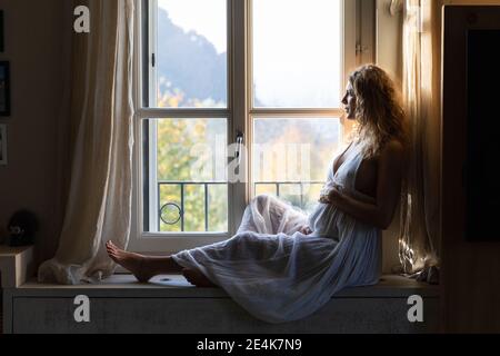 Donna incinta premurosa che tocca il ventre mentre si guarda attraverso la finestra a. casa Foto Stock