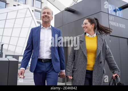 Donna d'affari sorridente e uomo d'affari che parlano mentre camminando contro l'ascensore a. fermata dell'autobus in città Foto Stock