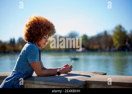 Donna mid adult con capelli crespi utilizzando il telefono cellulare a Estanque Grande del Retiro stagno il giorno di sole, Spagna Foto Stock