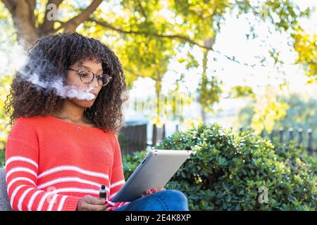 Giovane donna che usa il tablet digitale mentre fuma la sigaretta elettronica a. parco pubblico Foto Stock