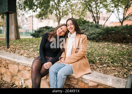 Madre sorridente con la testa sulla spalla della figlia seduta su trattenuta muro nel parco Foto Stock