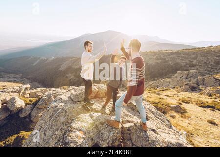 Amici allegri che si danno high-five l'uno all'altro mentre si levano in piedi sopra montagna durante il giorno di sole Foto Stock