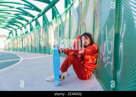 Giovane donna che tiene lo skateboard mentre si siede con la mano in capelli sul ponte Foto Stock