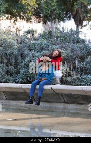 Sorridente giovane donna che copre gli occhi della fidanzata mentre si siede sopra muro di contenimento nel parco pubblico Foto Stock