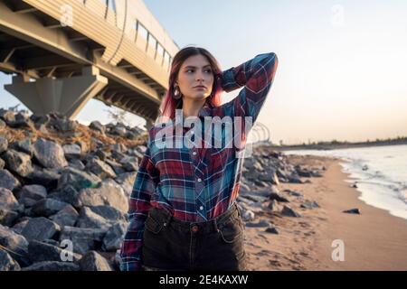 Femmina Hipster con la mano in capelli guardando lontano mentre si è in piedi in spiaggia Foto Stock