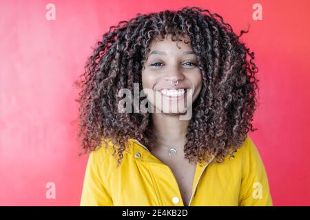 Donna sorridente con capelli ricci sorridente mentre si trova contro la parete rosa Foto Stock