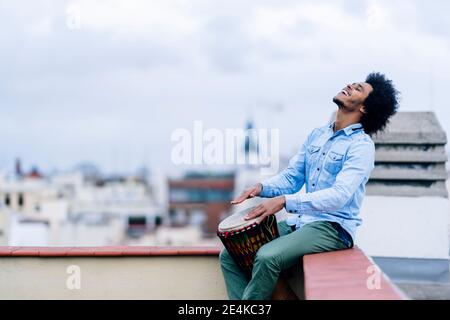 Felice giovane uomo che suona il tamburo mentre si siede sulla terrazza contro il cielo Foto Stock