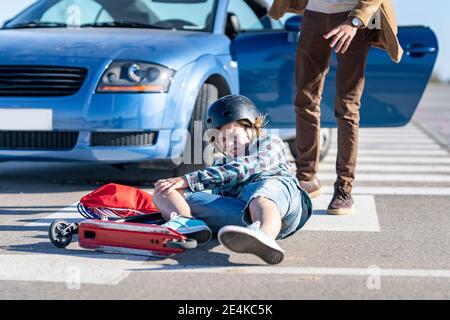 Ragazzo che cade dallo scooter a spinta con l'uomo in piedi auto su strada dopo un incidente Foto Stock