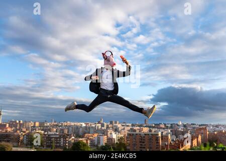 Uomo d'affari che indossa maschera di maiale saltando mentre prendendo selfie in città contro il cielo Foto Stock