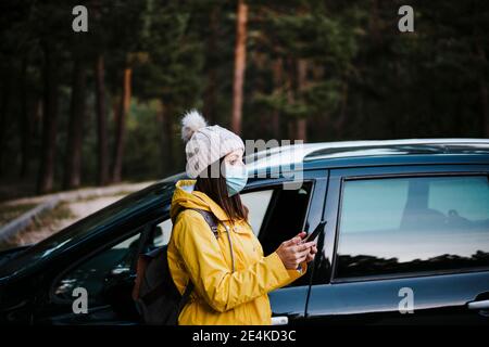Donna che indossa un impermeabile giallo e una maschera facciale in piedi vicino all'auto, tenendo in mano uno smartphone Foto Stock