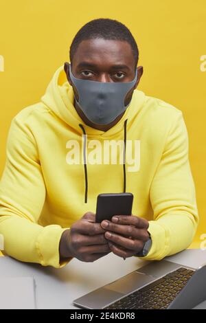 Ritratto verticale dell'uomo afroamericano contemporaneo che indossa la maschera mentre si siede alla scrivania su sfondo giallo pop Foto Stock
