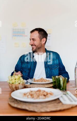 Buon professionista maschile avendo cibo a tavola mentre si guarda via in soggiorno Foto Stock