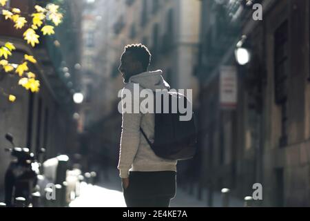 Giovane uomo con zaino che guarda lontano mentre si sta in piedi sulla strada in città Foto Stock