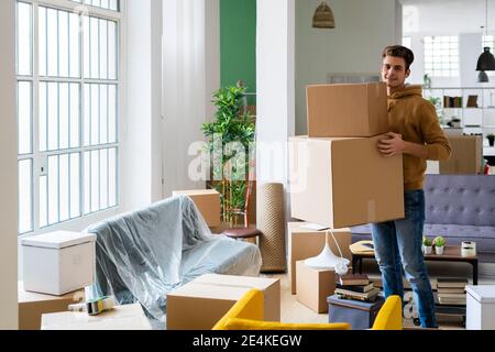Sorridente giovane uomo che porta scatole di cartone in soggiorno di nuovo appartamento Foto Stock