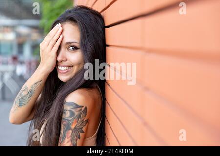 Giovane donna sorridente mentre ricopre il viso con la mano appoggiata parete Foto Stock