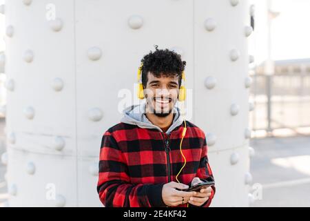 Uomo che indossa una camicia con cappuccio per ascoltare la musica attraverso le cuffie mentre si è in piedi con il telefono cellulare contro la parete Foto Stock