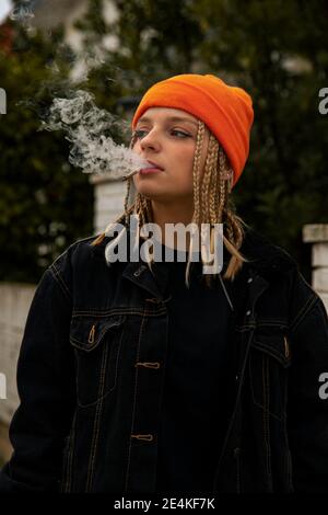 Donna alla moda che indossa una giacca nera che guarda lontano mentre fuma la sigaretta Foto Stock