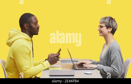 Vista laterale ritratto di due uomini d'affari contemporanei seduti di fronte l'uno all'altro alla scrivania durante il meting e sorridendo contro il giallo sfondo Foto Stock