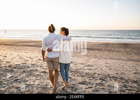 Giovane coppia con le braccia intorno a camminare in spiaggia durante il tramonto Foto Stock