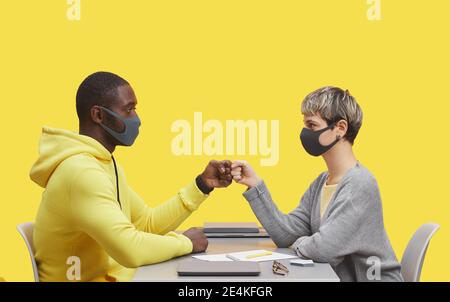 Vista laterale ritratto di due giovani uomini d'affari che indossano maschere e colpi di pugni mentre si siedono uno di fronte all'altro alla scrivania durante il meting contro il giallo Foto Stock