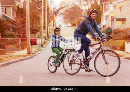 Ritratto in bici per tutta la famiglia con un piccolo ragazzo felice la bicicletta tandem di traino è collegata al padre sulla strada urbana Foto Stock